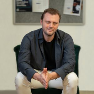 Ein Mann sitzt auf einem Hocker, stützt. die Ellenbogen auf die Knie und schaut in die Kamera