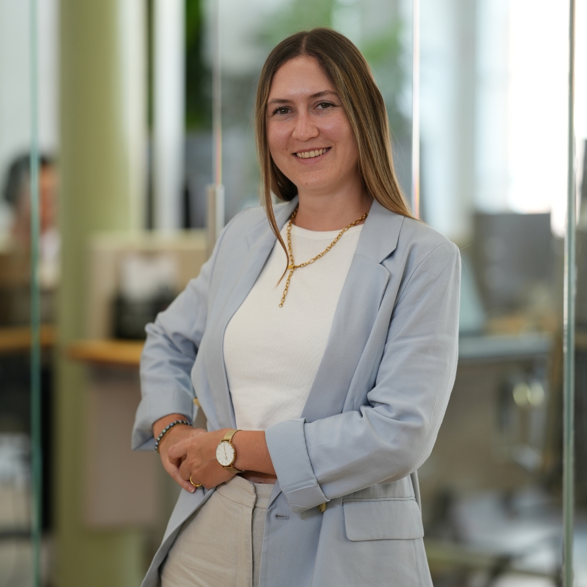 Eine junge Frau steht mit einem Blazer in einem Büro und lacht in die Kamera